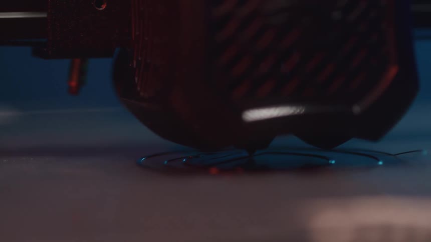 A closeup shot shows a 3d printer nozzle extruding a thin filament to build a circular object layer by layer on a flat surface