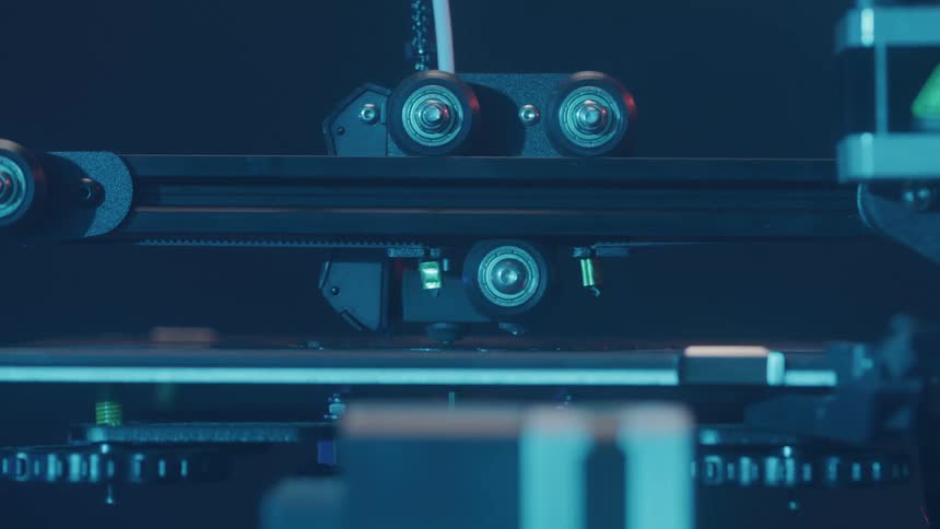 A closeup shot shows the mechanical gantry and rollers of a 3d printer moving during the printing process lit by a cool blue light