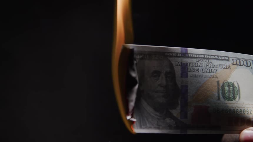 A 100 bill is held between a persons fingers as it slowly burns in a dark room