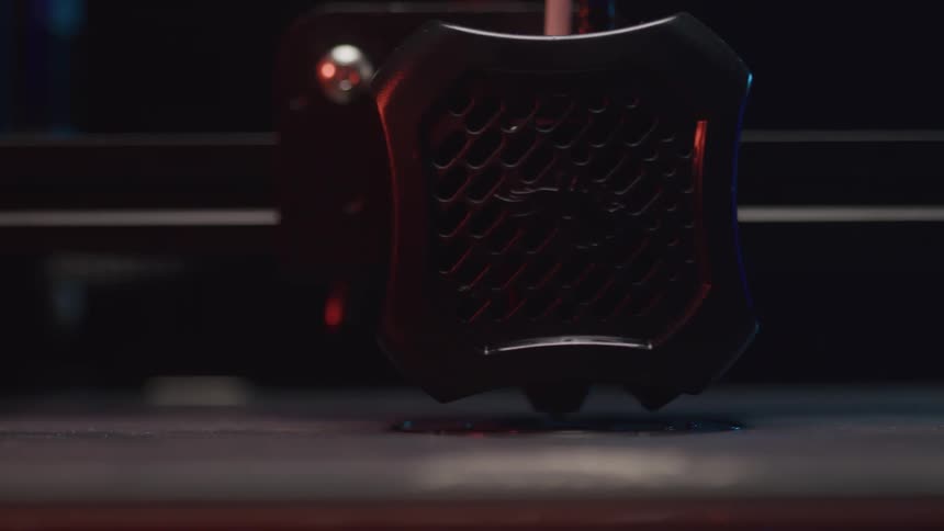 A closeup shot captures a 3d printers nozzle moving back and forth building a dark object layer by layer on the print bed
