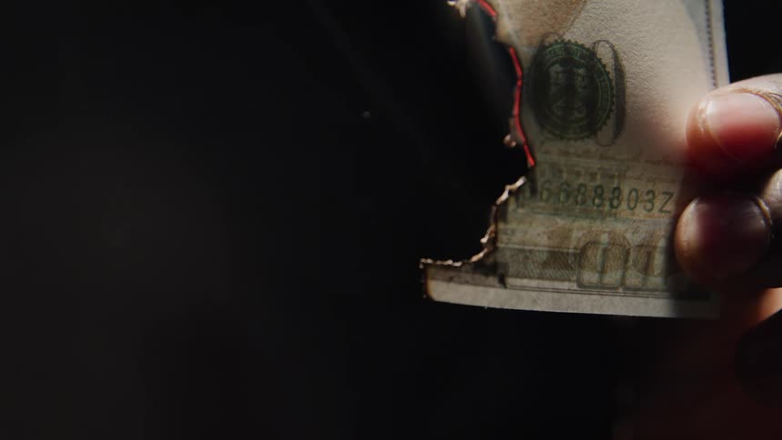 A closeup shot shows a hand holding a one hundred dollar bill as it smolders and turns to ash