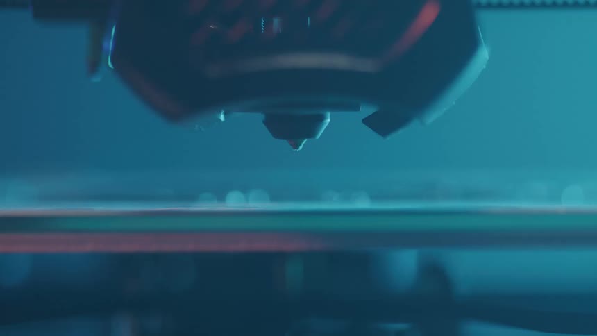 A 3d printer nozzle moves across a build plate extruding glowing material to form layers of an object the closeup view highlights the intricate and precise process of additive manufacturing under blue light