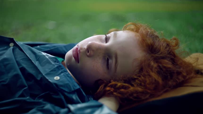 A young person with striking red curly hair lies on what appears to be a lush green lawn gazing upwards their relaxed posture and the natural surroundings suggest a moment of quiet contemplation or enjoyment of the outdoors