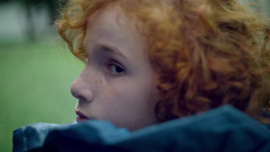 Redheaded girl wearing a raincoat looks over her shoulder while outdoors in a park
