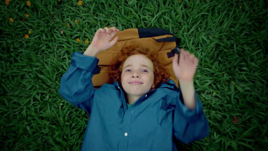 A relaxed boy with red curly hair lies on his backpack in the green grass smiling and raising his hands