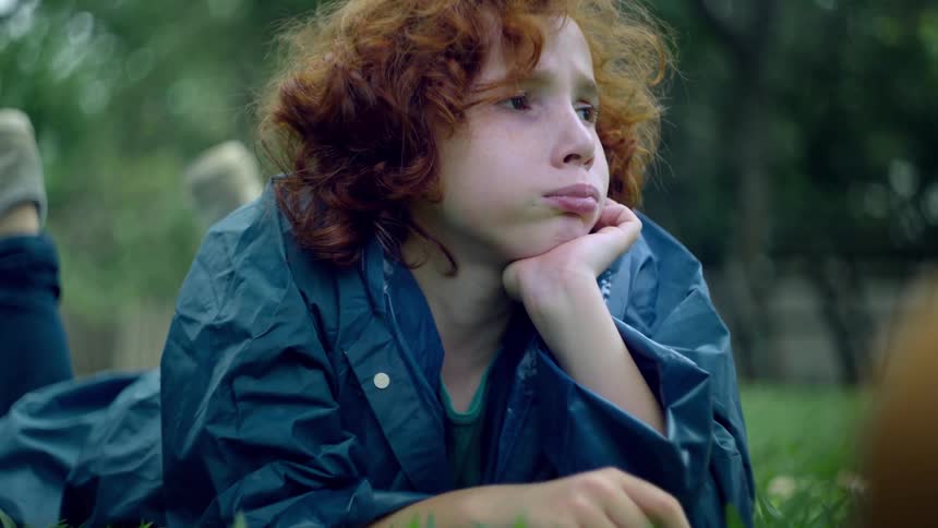 A person with curly red hair lies on their stomach in a field of green grass resting their chin on their hands while looking off into the distance their contemplative expression and the natural setting evoke a sense of peaceful introspection