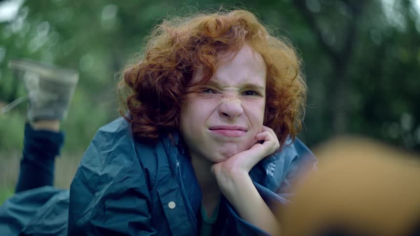 Child with red hair squints playfully while lying on the grass