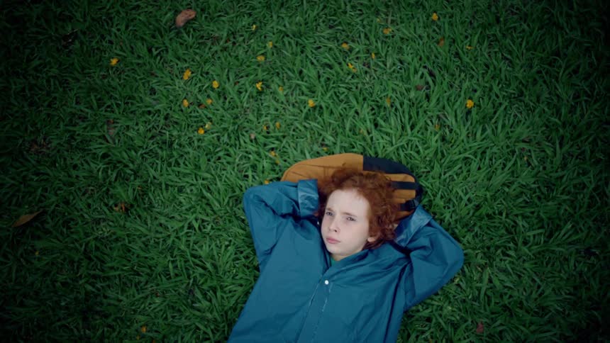A young person with curly reddishbrown hair dressed in a blue jacket lies comfortably on a vibrant green lawn with small yellow flowers scattered across it they are looking directly up at the camera with their hands behind their head
