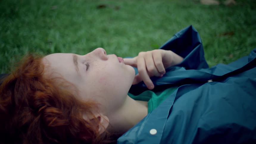 A redhaired child wearing a blue shirt lies on the green lawn grass at midday resting their index finger on the side of their face in a thoughtful pose