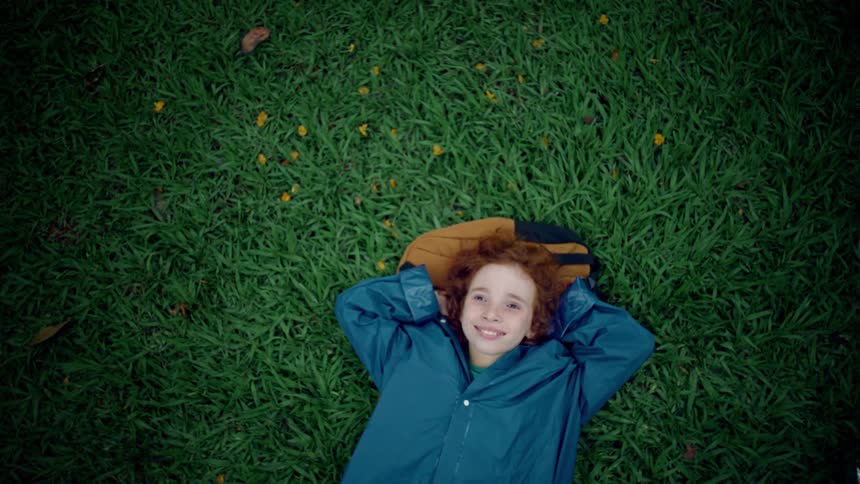 Smiling red haired boy in a blue jacket laying on his back on the lawn