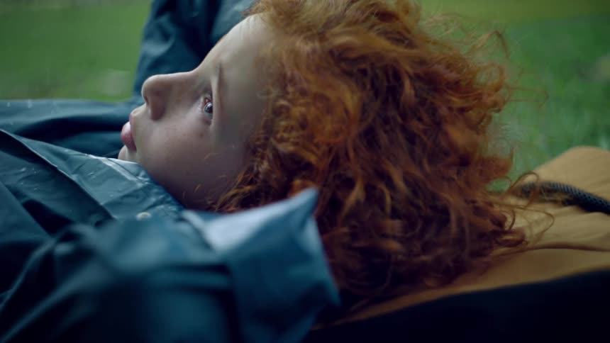 A child with curly red hair lies on their side on a brown backpack with lush green grass visible around them they are dressed in a blue jacket and appear to be resting or contemplating while looking up