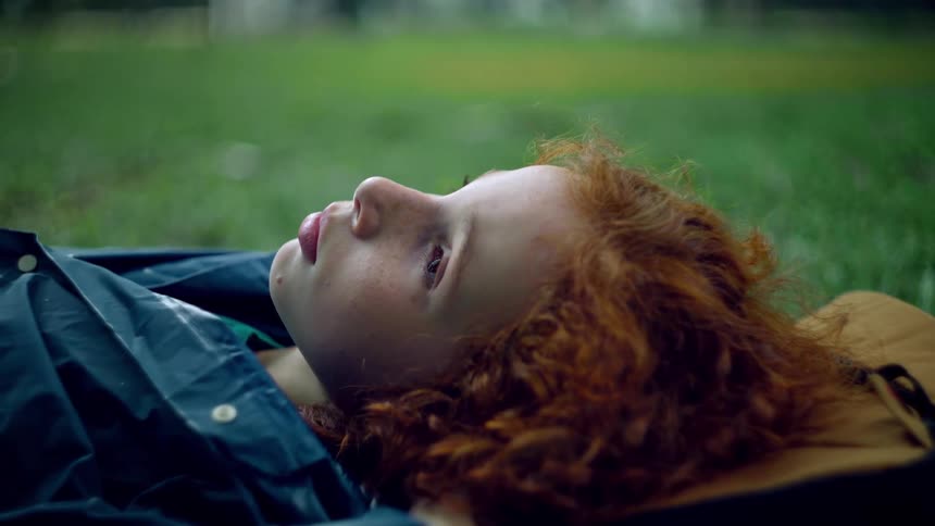 A young person with striking red curly hair lies on their back in a grassy area looking upwards with a contemplative expression they are dressed in a dark jacket seemingly at ease amidst the natural surroundings
