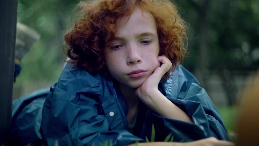 A child with curly red hair wearing a blue raincoat lying on green grass they appear to be resting or observing something with a contemplative expression occasionally closing their eyes