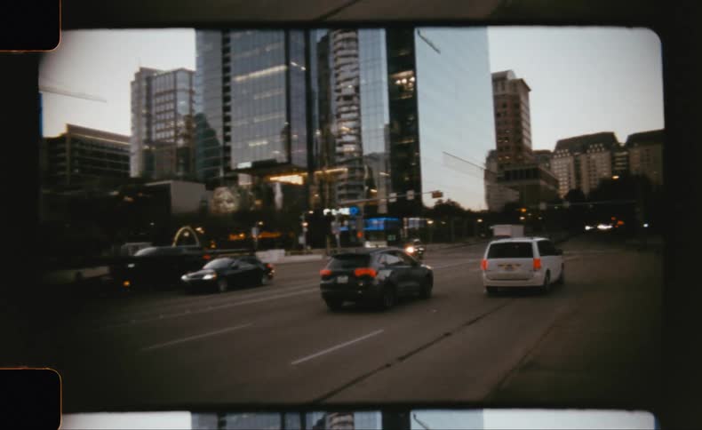 Traffic moves through a downtown street lined with illuminated skyscrapers at dusk