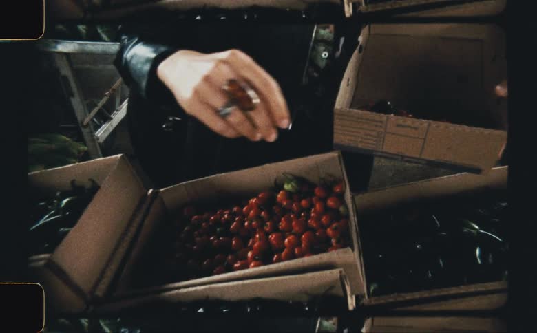 A pair of hands wearing a large ring is seen sifting through boxes filled with various types of produce including what appears to be red cherries and possibly other darker fruits or vegetables the scene is framed with rounded vignettelike corners