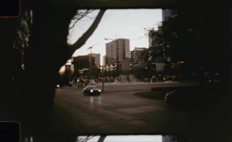 A car drives along a city street at dusk its headlights cutting through the dimming light in the background tall buildings and what appears to be a fountain are visible hinting at an urban landscape transitioning from day to night
