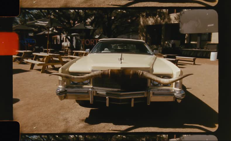 A vintage car is parked directly in front of outdoor picnic tables in a sunlit setting