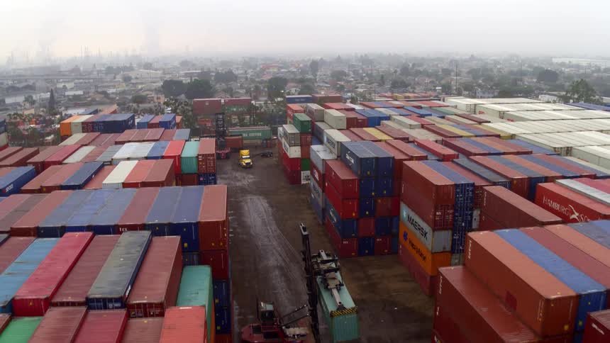 A container port with a large area filled with stacked containers as forklifts stack them and trucks move containers in and out at midday