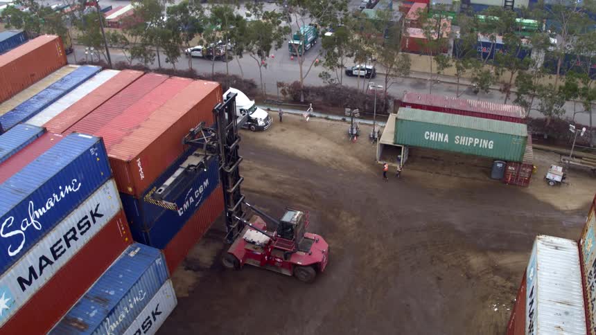 Workers walk around a business site while a heavy vehicle moves to grab a shipping container