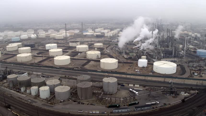 An aerial view shows a vast oil refinery with smoke billowing from stacks surrounded by storage tanks a highway and railroad tracks