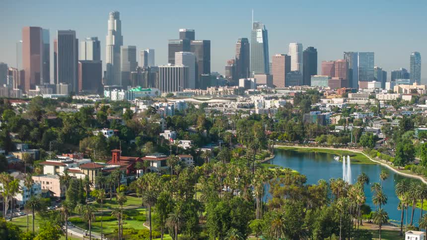 A cityscape shows downtown los angeles with a lush park and lake in the foreground