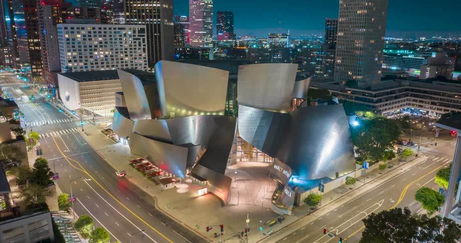 An aerial shot at dusk or night captures the walt disney concert hall in downtown los angeles with its distinctive curved metallic architecture glowing against the city lights traffic flows around the illuminated building and surrounding streets