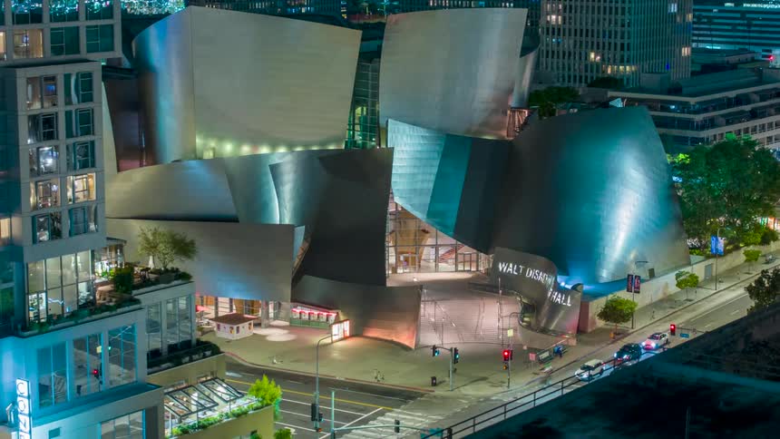 An aerial shot at night reveals the architectural marvel of the walt disney concert hall with its distinctive curved metallic facade brightly lit surrounding it the citys urban landscape glows with the soft lights of buildings and passing cars