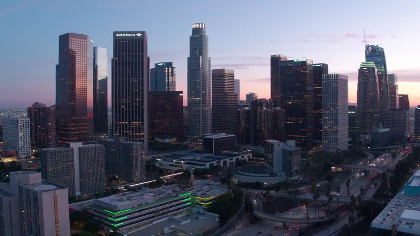 An aerial view showcases a sprawling Los Angeles skyline at dusk with numerous skyscrapers rising against a fading light below the intricate network of city streets and highways is visible bustling with the glow of vehicle lights