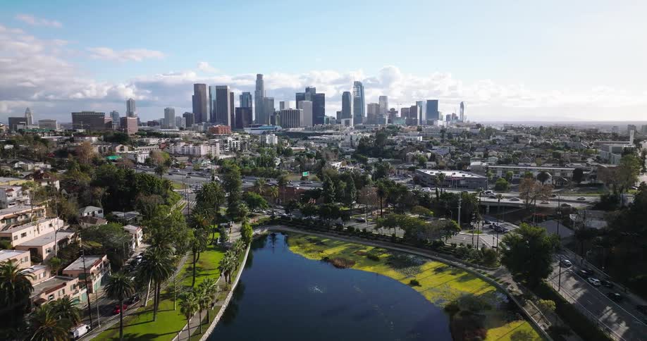 An aerial shot flies forward over a city park and lake moving towards the downtown los angeles skyline on a partly cloudy day