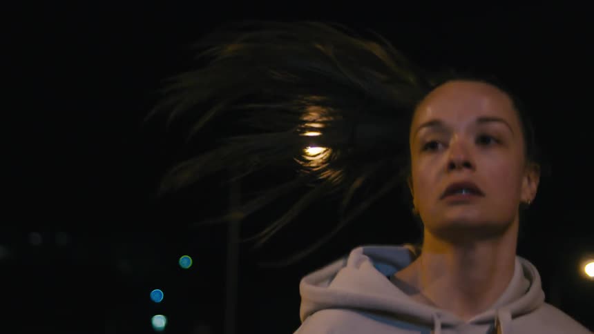 A woman with dark hair tied back wearing a lightcolored hoodie running at night the background is dark with outoffocus streetlights creating bokeh effects