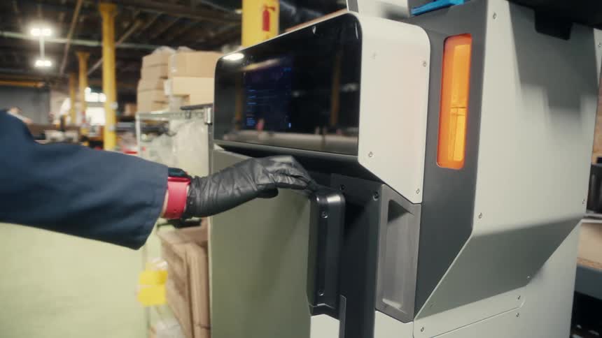 A person wearing dark gloves operating a sophisticated 3d printer they meticulously remove a component from the machine and then close its clear front door indicating the completion or a phase of a printing task in an industrial setting