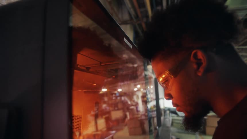 A man in safety glasses and black gloves inspects a glowing orangelit machine