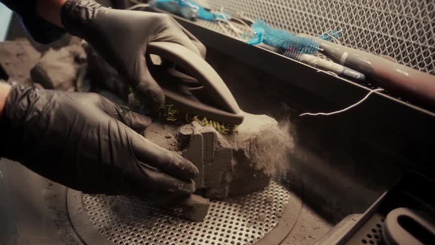 A person in black gloves uses a wire brush to clean a metal object on a mesh surface with fine detailing tools close at hand