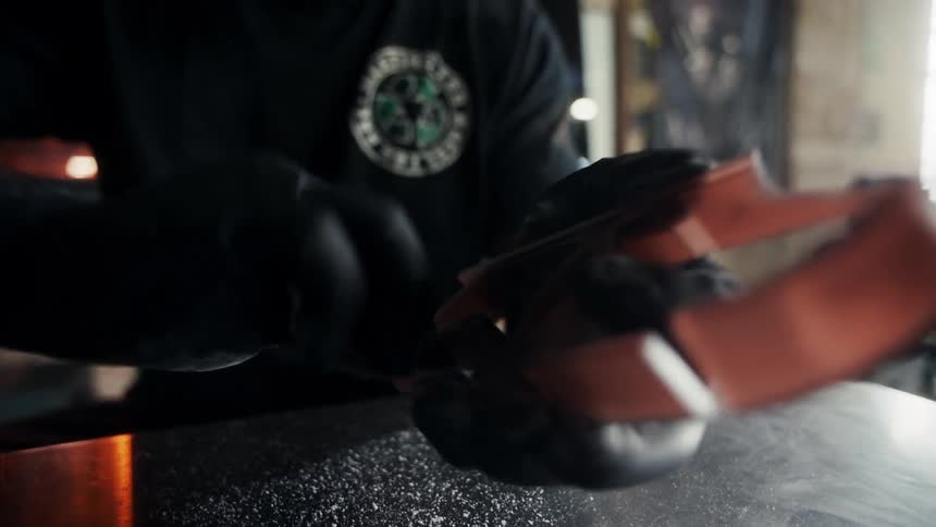 A craftsman wearing gloves is polishing a leather shoe sole in a workshop