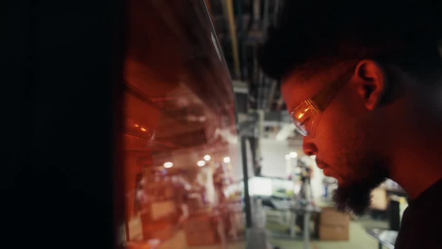 A person leans over a large reflective panel in a cluttered industrial space with metal equipment and stacked boxes
