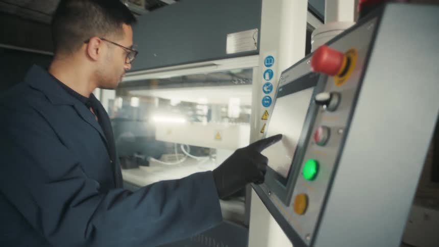 A person wearing gloves operates a touchscreen control panel of industrial machinery with various buttons and indicators