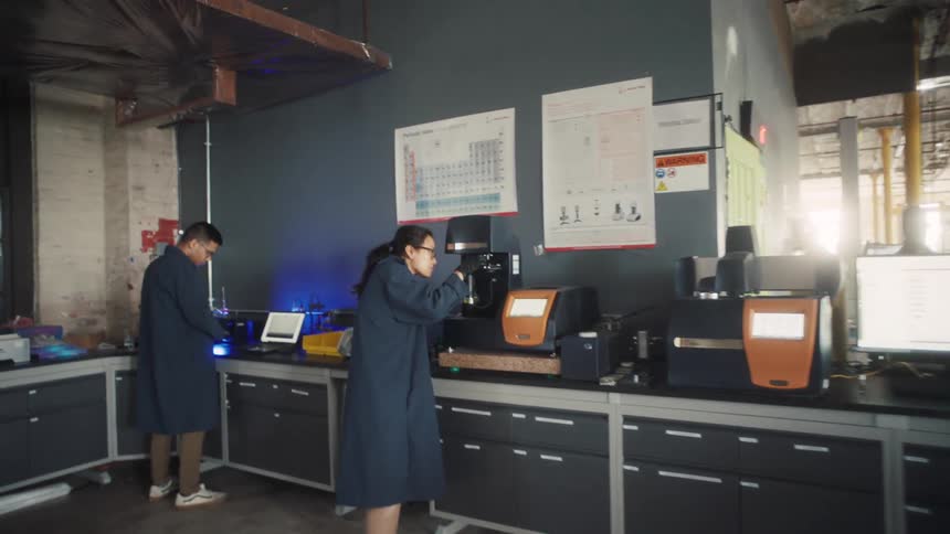 Two people wearing blue lab coats work at individual stations along a long laboratory counter filled with specialized equipment