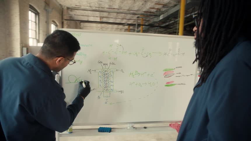 Male worker writes on a white board while explaining chemistry to his fellow black worker in a lab factory during daytime
