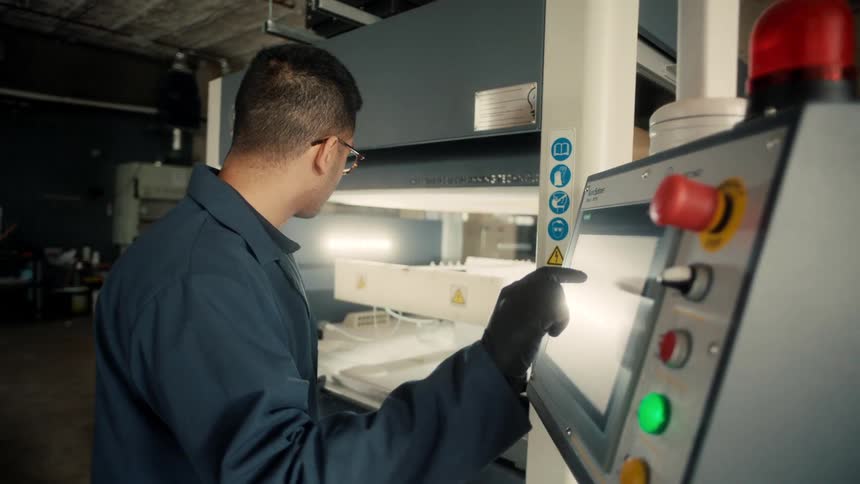 A person in a blue lab coat and black gloves operates a control panel with lit buttons and indicators in an industrial workspace