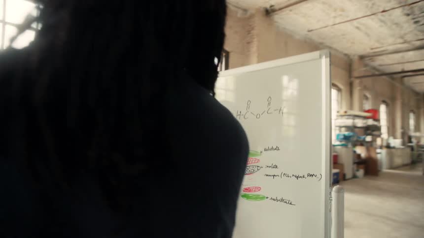 Two people in lab coats write on a whiteboard covered in technical diagrams and equations inside a spacious industrial lab