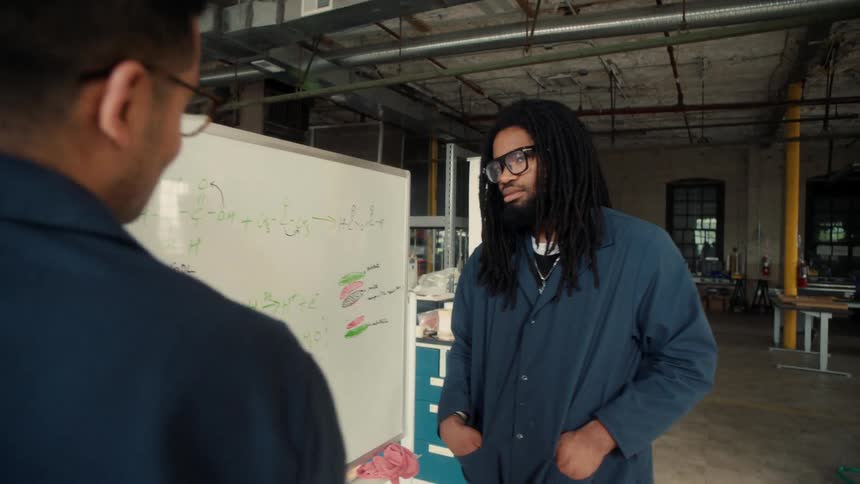Male worker explains chemistry to another black worker on a white board while standing a lab factory