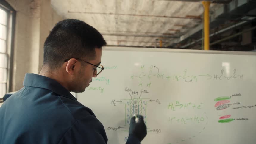 Male worker explains chemistry to another black male worker on a white board in a lab factory during daytime