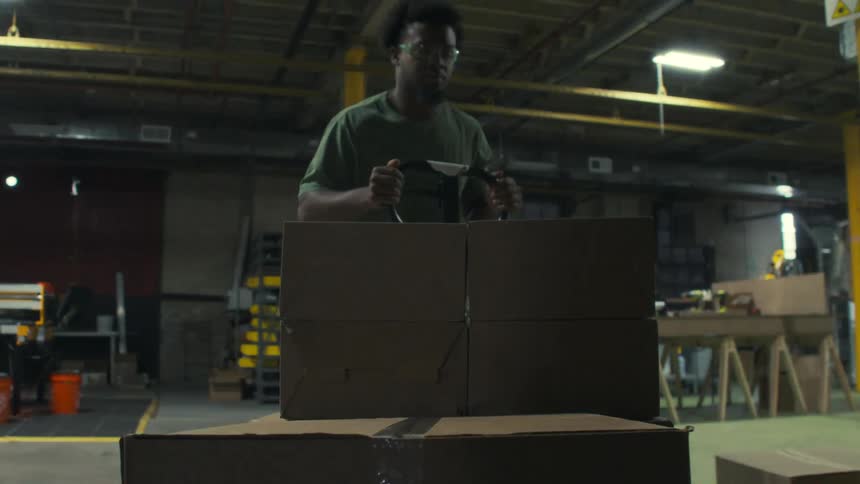 Young black worker arranges and packs boxes on a platform truck to move them around a factory during daytime