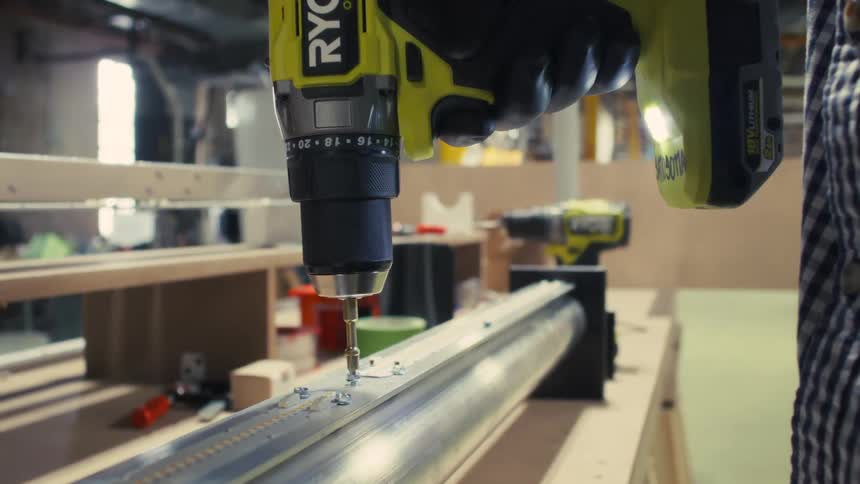 In a workshop a worker wearing a plaid shirt uses a power drill to drive a screw into a metal component on a workbench as someone walks by in the background