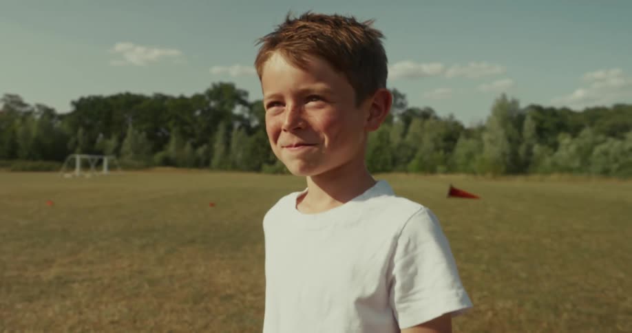 A child in a white tshirt stands in tall grass with trees and a clear blue sky in the background
