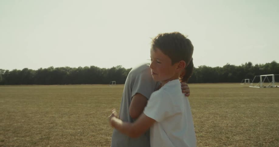 Two children stand on a grassy field clasping hands and hugging over a soccer ball facing each other at midfield
