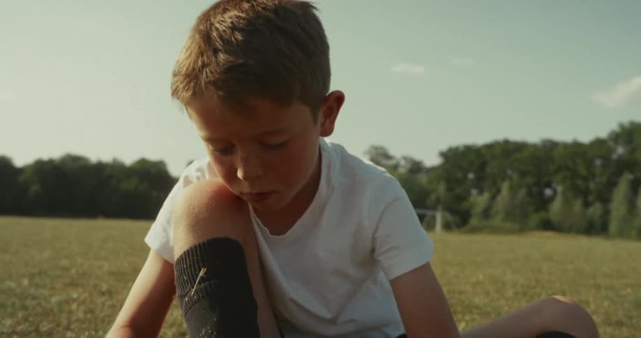 A child in a white shirt and black shorts is sitting on a grassy field tying the laces of black soccer cleats with a soccer goal visible in the background