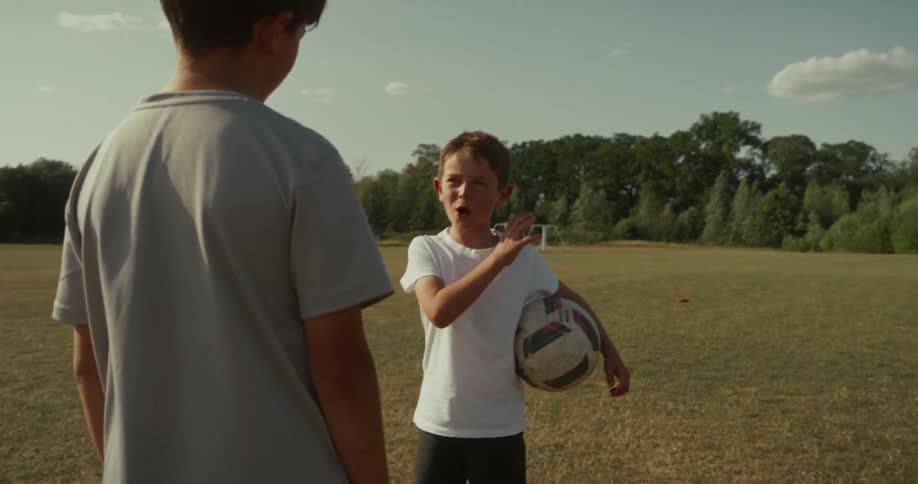 Two children stand on a grassy soccer field one holding a ball gesturing with goalposts in the distance