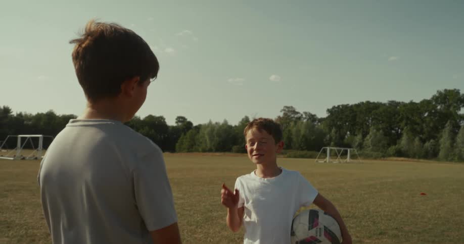 Two children stand on a grassy soccer field one holding a ball while gesturing with goalposts in the distance