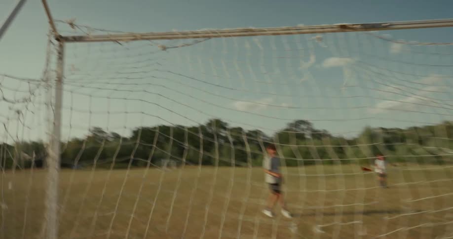 A person in a grey shirt and black shorts kicks a soccer ball towards a goal viewed through the net the ball soars over the goal and away from the field as the player stands in disappointment